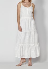 Vit maxiklänning med ögla broderi, tunna justerbara axelremmar, framknäppning och en lager-puffkant.