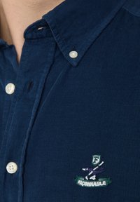 Camicia di velluto a coste blu navy con colletto classico, chiusura con bottoni bianchi e logo ricamato verde con un design di uccello sul petto.