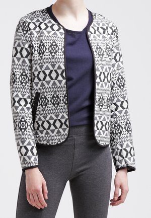 Vrouw draagt een zwart-witte jas met geometrisch patroon over een marineblauw shirt en donkergrijze broek, staand tegen een effen achtergrond.