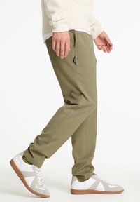 Olívazöld szűkülő nadrág oldalzsebekkel és egy kis fekete márkajelzéssel, fehér és szürke sportcipőkkel párosítva, amelyek gumitalppal rendelkeznek.