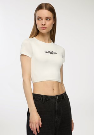 Top corto bianco realizzato in un tessuto liscio, con una stampa di una farfalla nera e un testo sul petto. Abbinato a jeans neri a vita alta.