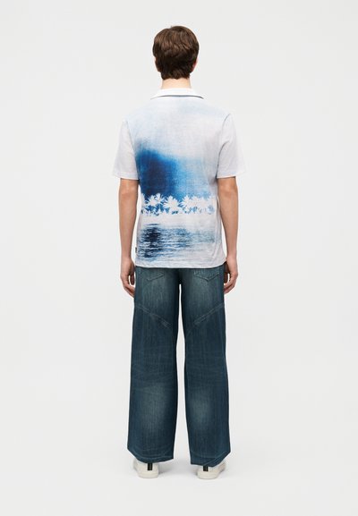 Νεαρό άτομο που φοράει λευκό κοντομάνικο πουκάμισο με μπλε σχέδιο από φοίνικες και νερό στην πλάτη, συνδυασμένο με μπλε τζιν παντελόνι φαρδιάς γραμμής και λευκά παπούτσια.