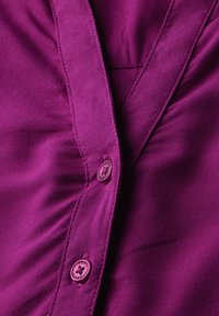 Camicia di seta viola con una texture liscia, dotata di un design a scollo a V e bottoni coordinati. Sono visibili pieghe notevoli e dettagli di cucitura fine.