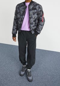 Giubbotto bomber imbottito in camouflage nero e grigio, maglietta viola a girocollo, pantaloni jogger neri con logo Nike e sneakers grigie.