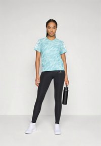 Ženska stoji v črnih leginsih, svetlo modri športni majici, belih supergah, v roki drži črn plastenko za vodo proti beli ozadju.