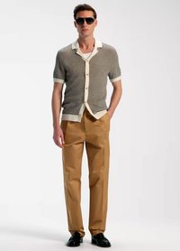 Cardigan lavorato a maglia a maniche corte in beige e crema, con bottoni sul davanti; abbinato a pantaloni sartoriali color marrone chiaro e scarpe nere; il modello indossa occhiali da sole.