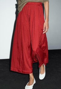Pantalons larges rouges avec des plis, en tissu léger. Portés avec des chaussures blanches à enfiler, arborant une texture lisse et une couleur riche.
