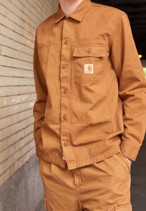 Person trägt ein braunes Hemd mit Knöpfen und passende Hose mit einem Carhartt-Logo-Patch auf der Brusttasche, steht vor einer Backsteinmauer.