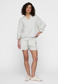 Felpa in misto cotone grigio chiaro con scollatura a V e maniche oversize, abbinata a pantaloni corti coordinati con vita regolabile tramite coulisse. Sneakers bianche.