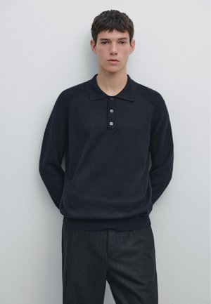 Mladý muž s krátkými tmavými vlasy, oblečený do tmavě modrého polotrika s dlouhým rukávem a tmavých kalhot s proužky, stojící u světle zbarvené zdi.