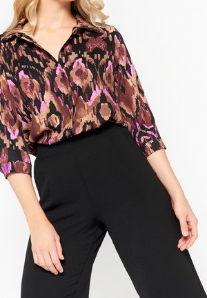 Frau trägt eine braun-schwarz-pink gemusterte Bluse mit dreiviertelÄrmeln, die in eine hoch taillierte schwarze Hose gesteckt ist.