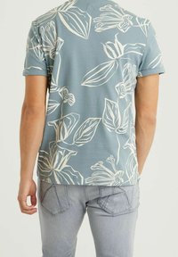 Lyhythihainen sininen T-paita valkoisella kukkakuviolla. Pehmeä kangas, rento istuvuus, pyöreä kaula-aukko, näkyviä saumoja takana. Yhdistetty vaaleanharmaisiin farkkuihin.