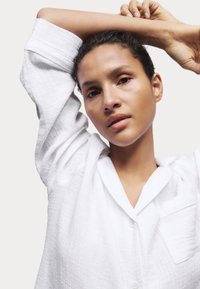 Chemise blanche texturée à manches longues, avec col et poche ; manches retroussées ; tissu léger et coupe décontractée.