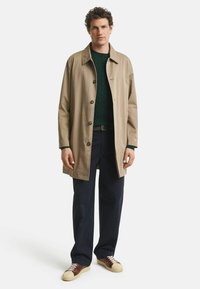 Beige Trenchcoat aus Baumwolle mit einer Knopfleiste vorne, seitlichen Taschen und einem lässigen Schnitt, kombiniert mit einem dunkelgrünen Pullover und navyfarbenen Hosen.