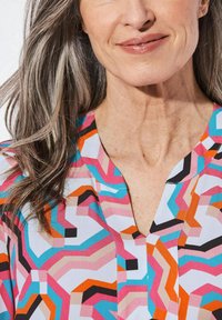 Bunte Bluse mit geometrischem Muster in Pink, Orange, Blau und Schwarz. Leichter Stoff mit V-Ausschnitt und kurzen Ärmeln.