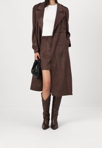 Brązowy zamszowy płaszcz z dużymi klapami, zapinany na podwójne guziki, z długimi rękawami. W połączeniu z dopasowaną brązową mini spódniczką i kowbojkami.