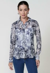 Una donna si erge indossando una camicia a fiori con bottoni e jeans blu, mostrando un'espressione sicura di sé su uno sfondo chiaro.