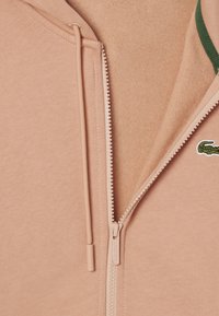 Sudadera con cremallera de color melocotón, hecha de material suave, con un acento verde y un parche de logo. Incluye una cremallera suave y cordones.