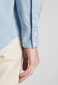 Main droite et poignet d'une personne portant une chemise bleu clair à manches longues avec un poignet boutonné et un pantalon couleur crème sur un fond uni.