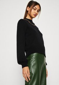 Black knitted sweater with puffed sleeves and ribbed hem, paired with shiny green faux leather pants. Gold chain necklace adds detail.