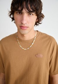 Braunes Baumwoll-T-Shirt mit einem kleinen, strukturierten Logo auf der linken Brust. Getragen mit einer gemusterten, metallischen Halskette. Lässige Passform.