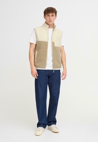 Beige Fleece-Weste mit hohem Kragen und Fronttaschen, kombiniert mit dunkelblauen Jeans und hellen Sneakers.