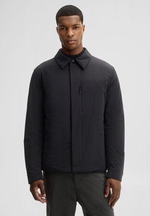 Mann trägt schwarze Jacke mit Reißverschluss und Kragen und graue Hose vor einfachem hellem Hintergrund.
