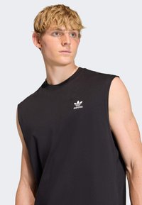 Čierne tielkov�é tričko vyrobené z jemnej bavlnenej látky. Na ľavej strane na hrudi je biely nápis Adidas. Minimalistický dizajn, pravidelný strih.