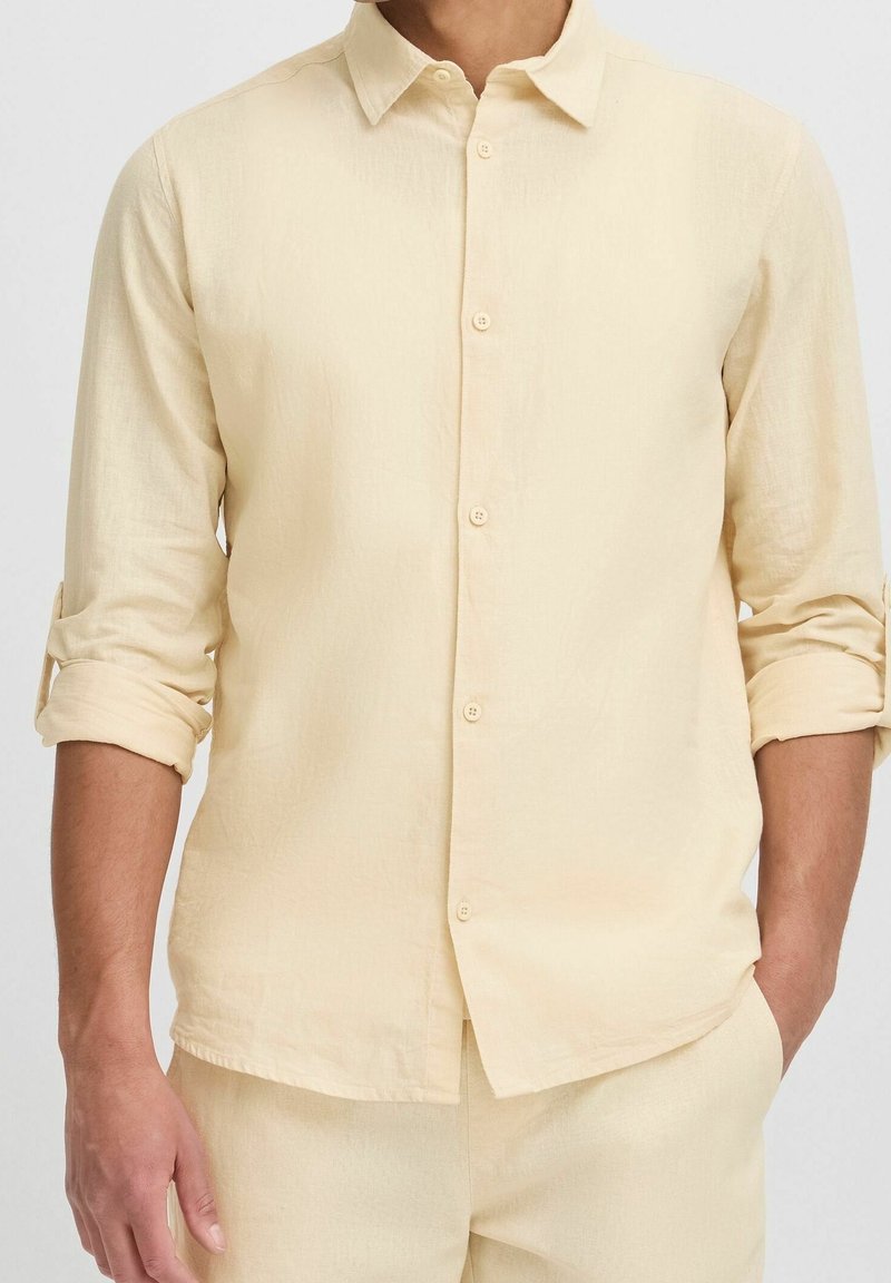 Homme portant une chemise beige à manches longues boutonnée avec les manches retroussées, assortie à un pantalon beige, une main dans la poche.