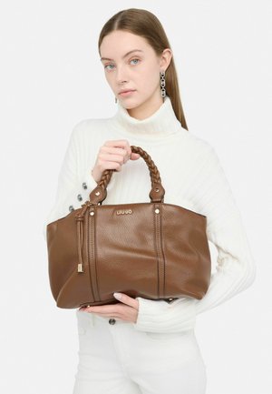Bruine leren totebag met gevlochten handvatten, gouden accenten en een gestructureerde afwerking. Model houdt het tegen een witte achtergrond.
