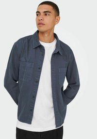 Camicia azzurra scuro con bottoni, dotata di due tasche sul petto e colletto, indossata sopra una maglietta bianca, mostrata su uno sfondo chiaro.