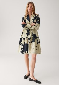 Eine Frau steht mit verschränkten Armen in einem knielangen, langärmeligen Kleid mit großen schwarzen und beigen Blumenmustern sowie schwarzen flachen Schuhen.