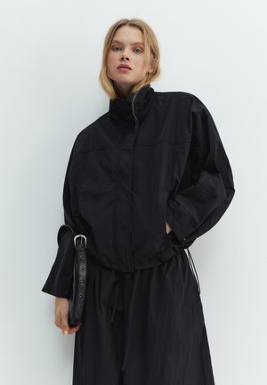 Blondynka ubrana w luźną czarną kurtkę i spodnie, trzymająca czarny pasek, stojąca na tle jednolitego jasnego tła.