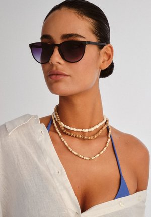Woman with slicked-back hair wears black sunglasses, layered shell necklaces, a blue bikini top, and a white button-up shirt draped on one shoulder.