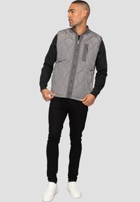 Gepolsterte graue Weste mit einem gesteppten Muster, Frontreißverschluss und einer Reißverschlusstasche; getragen über einem schwarzen Langarmshirt und schwarzen Hosen.