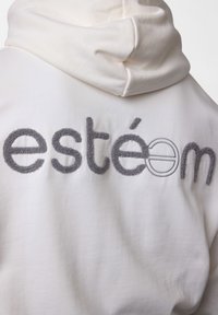 Haut à capuche blanc avec une texture douce, ornée de broderies grises "estéom" dans le dos. Détail de la capuche visible en haut.