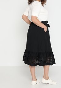 Svart knälång kjol gjord av lättviktsmaterial, med en volangsläp och intrikata utskärningar samt ett knytbälte fram.