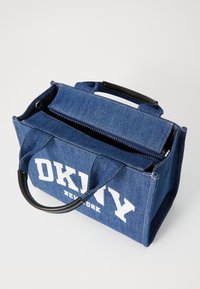 Sac fourre-tout en denim bleu avec des poignées en simili cuir noir, fermeture zippée et logo "DKNY" blanc imprimé sur le côté. Présente une forme rectangulaire.