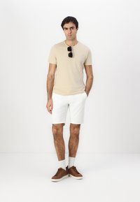Béžové tričko s krátkým rukávem, bílé šortky, hnědé sneakers a bílé ponožky. Černé sluneční brýle položené na tričku. Jednoduchý, neformální outfit.