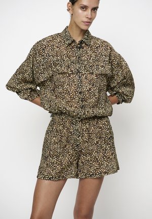 Vrouw draagt een bijpassend losse shirt en korte broek met luipaardprint, staand met de handen achter de rug tegen een effen witte achtergrond.