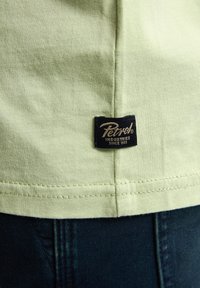 Camiseta de algodón verde claro con una etiqueta negra con texto dorado en la costura lateral inferior, con detalles de costura visibles sobre unos jeans de mezclilla oscuros.
