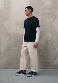 T-shirt noir à manches courtes superposé sur une chemise blanche à manches longues, associé à un pantalon large crème et des baskets multicolores.