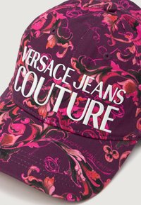 Căciulă purpurie detaliată cu modele florale roz și text alb "VERSACE JEANS COUTURE" în față. Dispune de o margine curbată și o construcție din material textil.