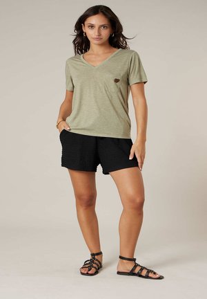 Femme debout portant un t-shirt à col en V vert olive clair avec une poche et un short noir texturé, associé à des sandales noires à brides.