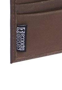 Cartera de cuero marrón con múltiples ranuras para tarjetas y un borde cosido; presenta una etiqueta de tela negra con texto blanco. Diseño compacto y minimalista.