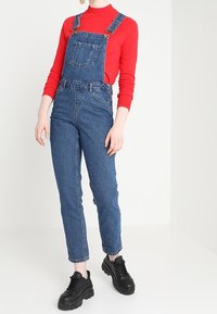 Salopette in denim con bretelle regolabili blu, tasca frontale e gambe con taglio affusolato, abbinata a una maglia rossa a maniche lunghe e sneakers nere chunky.