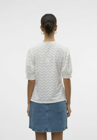 Hvid kortærmet top med et zigzag-mønster og øjenlignende detaljer, parret med en denim nederdel med to baglommer.