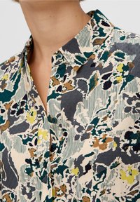 Blusa con colletto in un motivo floreale multicolore che include verdi, gialli e marroni, con chiusure a bottone e un tessuto strutturato.