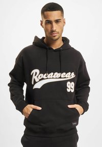 Fekete kapucnis pulóver pamutból, fehér "Rocawear 99" logóval. Elülső kenguruzsebe és állítható zsinóros kapucnija van.