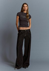 En ung kvinna står mot en grå bakgrund iförd en svart kortärmad t-shirt med text och vida mörk denimjeans samt silvriga spetsiga skor.